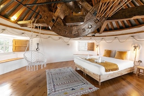 A bedroom with a wooden ceiling, a bed with white bedding, a hanging chair, and a patterned rug.