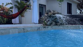 A swimming pool with a clear blue water surface, a white building with a tiled roof, and a hammock.