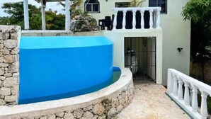 Una casa con pared de piedra, piscina azul, patio cubierto y techo de tejas rojas.