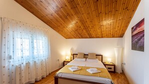 Une chambre avec un plafond en bois, un lit recouvert de draps blancs et agrémenté de deux oreillers, une table de chevet avec une lampe, et une fenêtre ornée de rideaux légers.