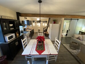 Ein Essbereich mit weißem Tisch und Stühlen, einem roten Läufer und einer roten Weihnachtsstern. Es gibt eine Küchenzeile mit Mikrowelle, Herd und Schränken. Im Hintergrund ist ein Kühlschrank zu sehen.
