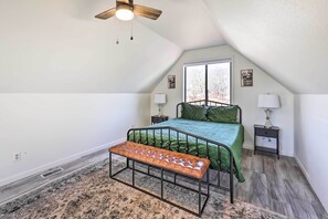 Un dormitorio con techo inclinado, cama con ropa de cama verde, dos lámparas de noche, un banco y una ventana con vistas a árboles.