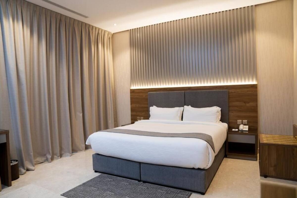 Une chambre d’hôtel moderne avec un grand lit, une tête de lit en bois, une table de chevet avec une lampe et une grande fenêtre avec des rideaux.