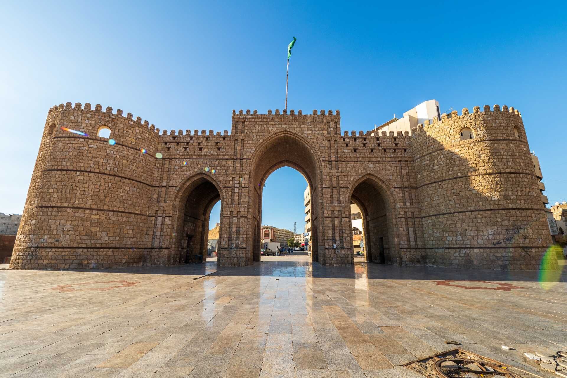 Un arco de piedra histórica con una bandera en la parte superior, que da paso a un área pavimentada.