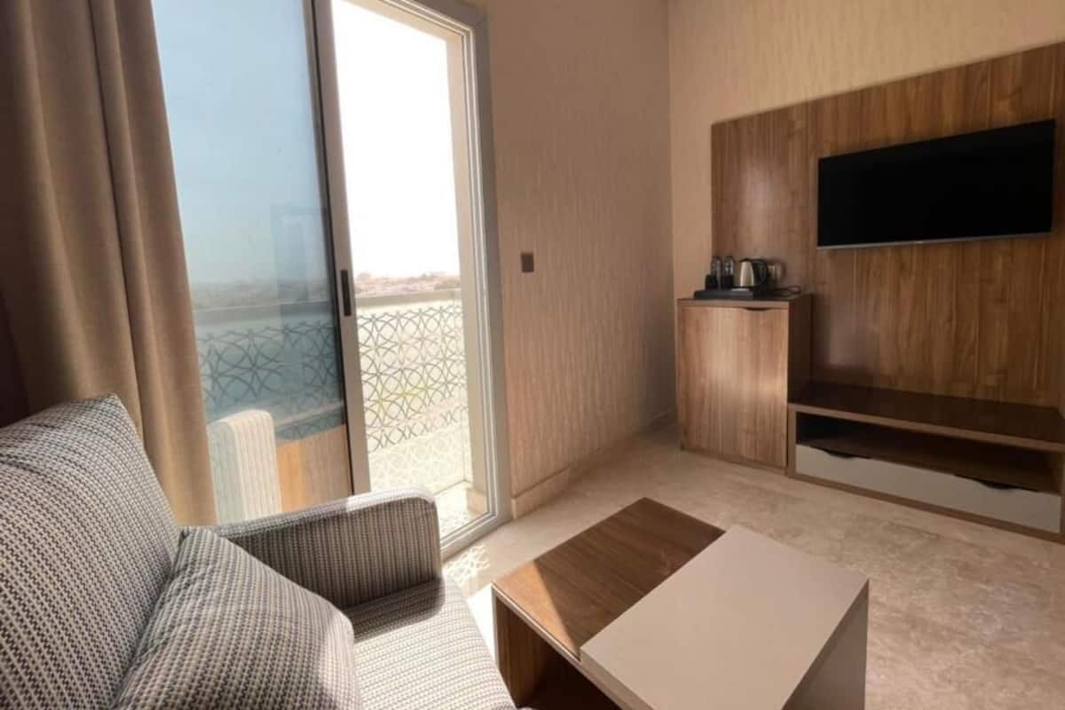 Une chambre d’hôtel moderne équipée d’un canapé, d’une table basse, d’un téléviseur fixé au mur et d’une porte-fenêtre coulissante donnant sur un balcon.
