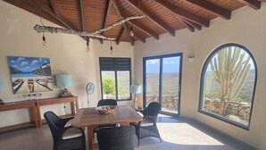 Una zona de menjador amb taula de fusta i cadires de vímet, un quadre a la paret i una finestra gran amb vista a un cactus.