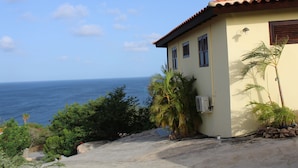 Una casa a la costa amb vista a l'oceà, amb teulada de rajola, un petit jardí amb palmeres i un camí d'accés de formigó.