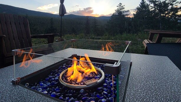 A square outdoor fire pit with a glass enclosure, surrounded by blue pebbles, set on a textured surface.