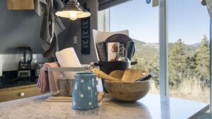 A kitchen with a marble countertop, a blue polka-dotted pitcher, a wooden bowl, and a window with a view of trees and hills.