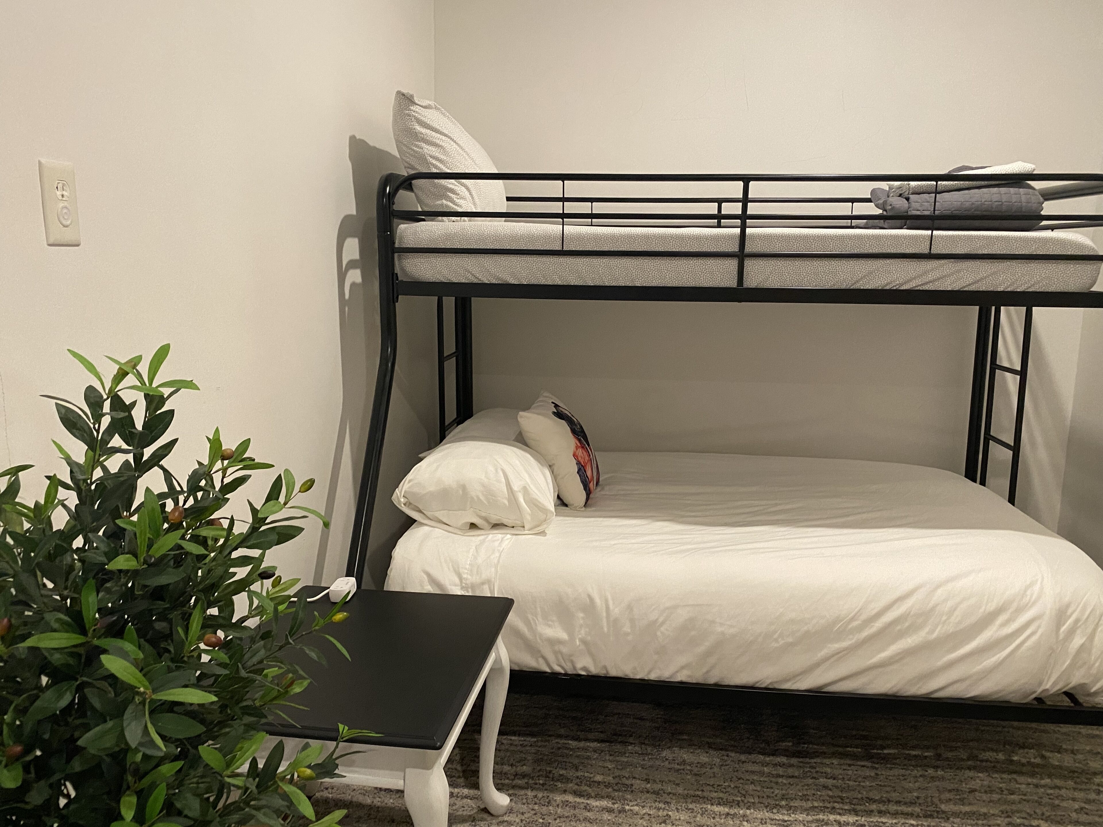 Una cama individual con ropa de cama blanca, una mesita pequeña y una planta en maceta en una habitación con paredes lisas.