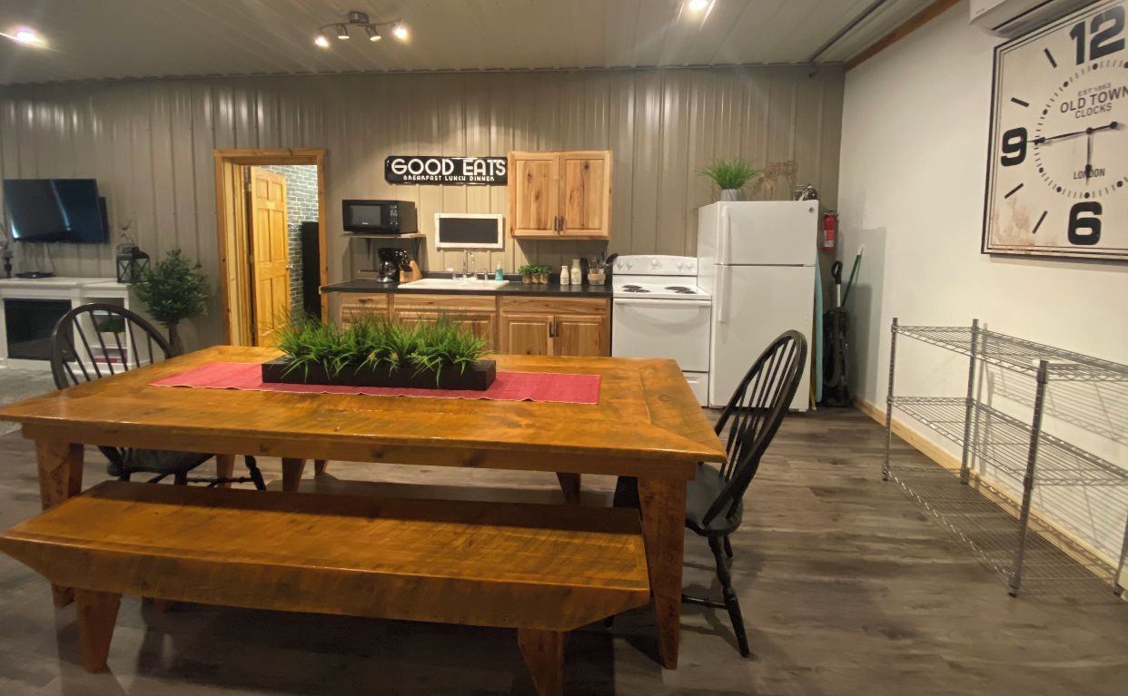 Un comedor con una mesa de madera, sillas y un banco. Se observa una cocina al fondo con armarios de madera, un microondas y un refrigerador. Un reloj está colgado en la pared.