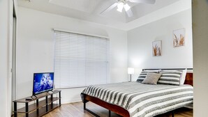 Un dormitorio con cama, televisor sobre un mueble, ventilador de techo y ventana con persianas.