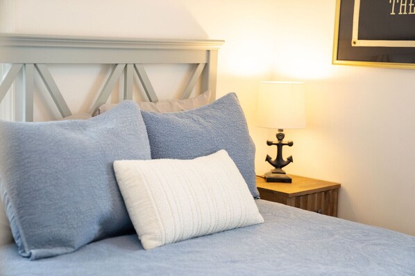 Una cama bien hecha con una disposición de almohadas azules y blancas, una mesita de noche de madera con una lámpara y un cuadro en la pared.