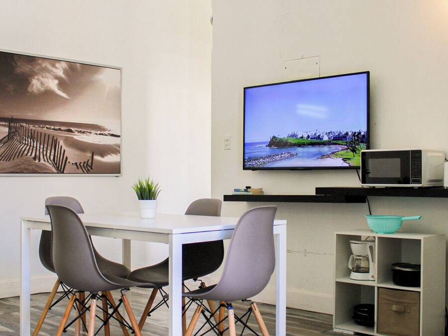 A modern room with a white table, four grey chairs, a flat-screen TV, a microwave, a coffee maker, and a shelf with books and a plant.