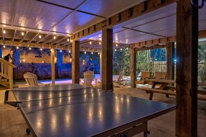 Un espace intérieur aménagé pour jouer au ping-pong, avec des piliers en bois, des guirlandes lumineuses et une piscine en arrière-plan.