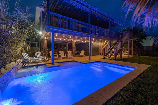 A backyard with a swimming pool, lounge chairs, and a covered patio area with string lights.