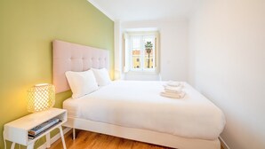 A neatly arranged bedroom with a large bed, a bedside table with a lamp, and a window offering a view of buildings and a palm tree.