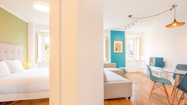 A modern hotel room with a bed, a sofa, a dining area with a table and chairs, and a TV.