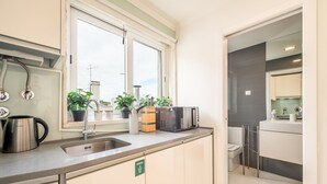 A compact kitchen with a sink, a kettle, and a microwave. There are potted plants on the windowsill and a green sign indicating a general waste bin.