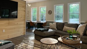 Ein modernes Wohnzimmer mit Ecksofa, Couchtisch und an einer Holzwand montiertem Fernseher. Große Fenster sorgen für natürliches Licht und bieten einen Blick ins Freie.