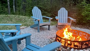 Ein Lagerfeuer, umringt von Adirondack-Stühlen, inmitten eines Waldes.