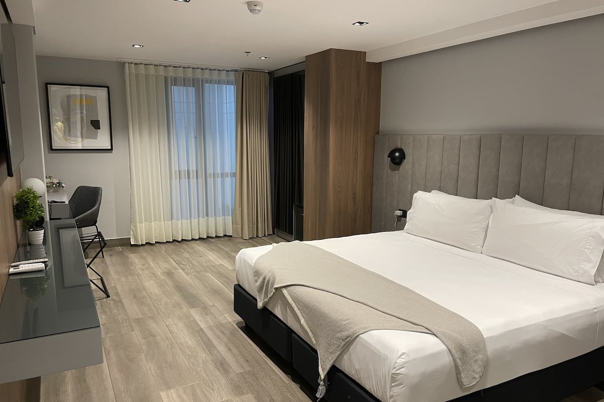 A modern hotel room with a large bed, a desk with a chair, a potted plant, and a window with curtains.