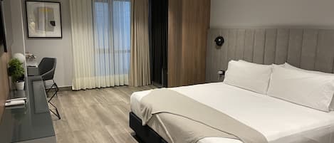 A modern hotel room with a large bed, a desk with a chair, a potted plant, and a window with curtains.