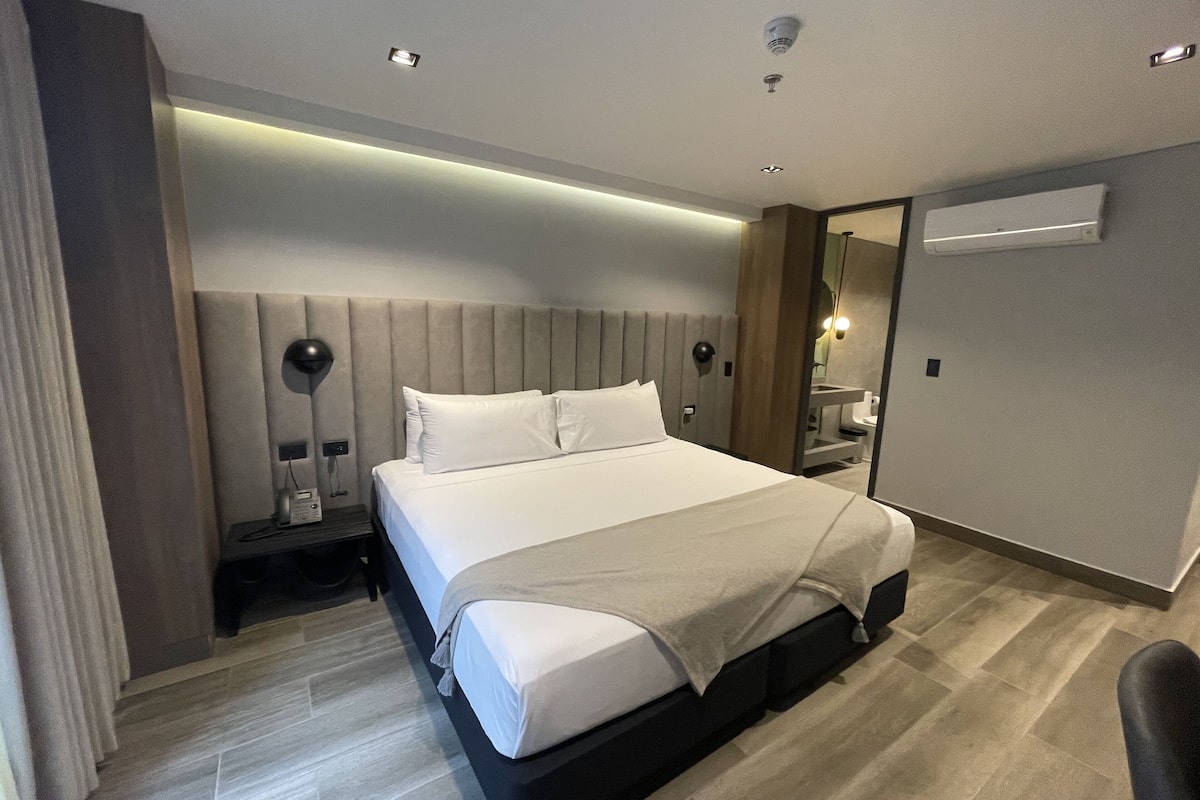 A modern hotel room with a large bed, bedside tables, a mirror, and a built-in air conditioning unit.