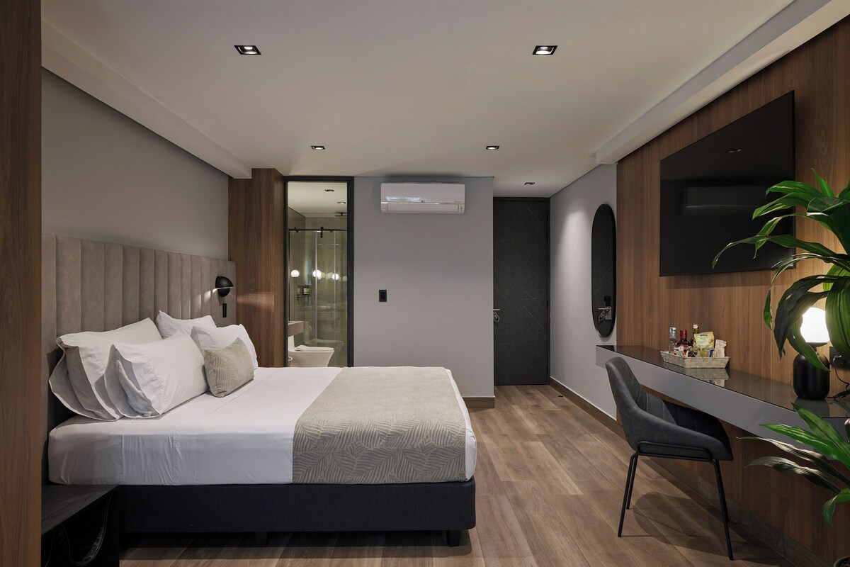 A modern hotel room with a bed, a desk, a chair, a TV, and an air conditioning unit.