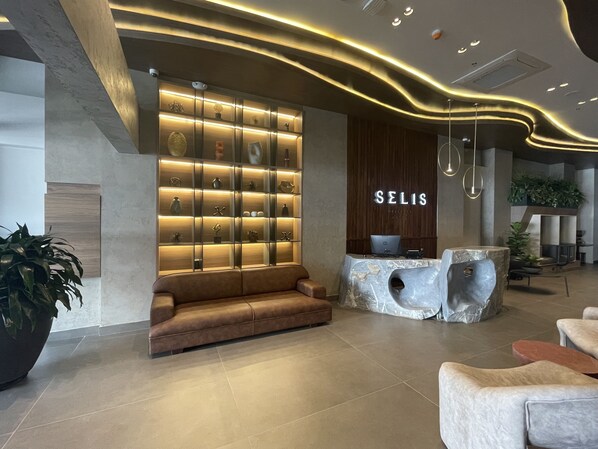 Una moderna zona de recepción de hotel con un mostrador de madera, un área de estar con un sofá marrón y una pared de estantes iluminados que exhiben diversos objetos.