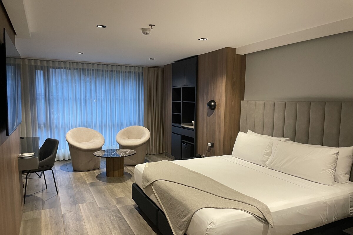 A modern hotel room with a large bed, two armchairs, a small table, a TV, and a wardrobe.