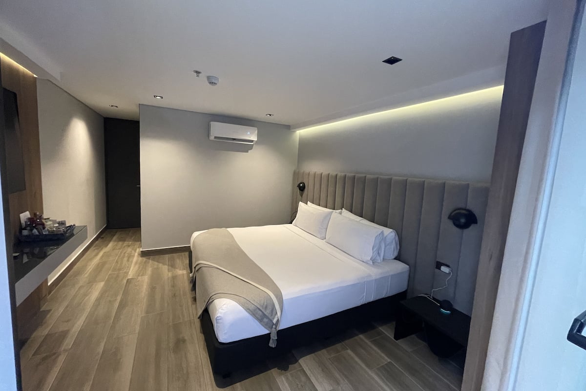 A modern hotel room with a large bed, a bedside table, an air conditioning unit, and a wooden floor.