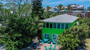 A green house with a dock extending into a green body of water, surrounded by trees and a clear sky. - Belle Vista (Madeira Beach)