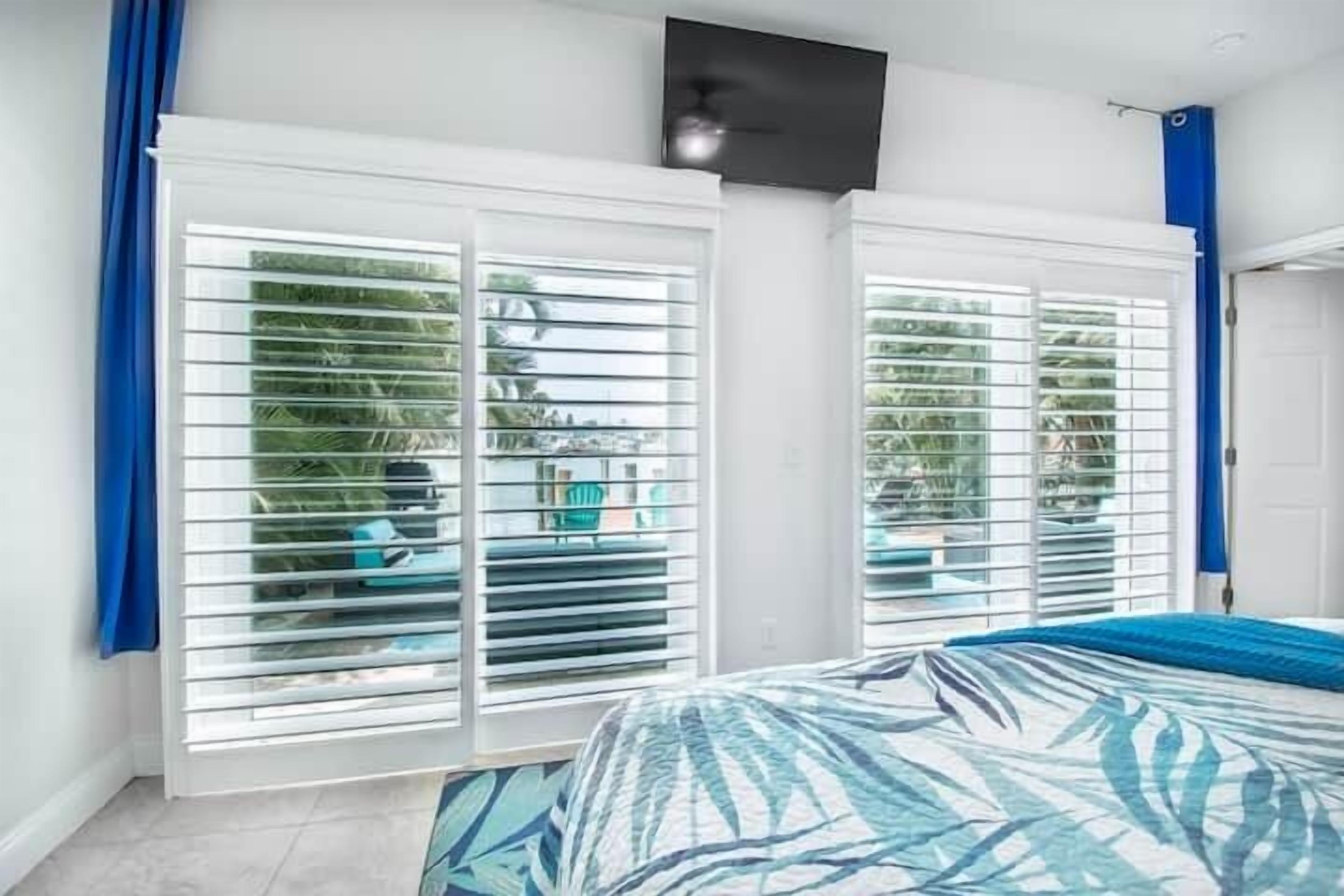A bedroom with a bed, white shutters, and a view of greenery outside.