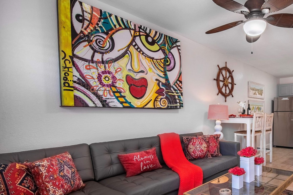 A living room with a gray sofa, red throw blanket, and patterned cushions. A colorful abstract painting is on the wall.