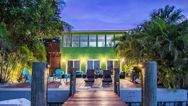 A wooden deck leading to a building with a green facade and large windows. There are outdoor seating areas with chairs and tables. - Belle Vista (Madeira Beach)