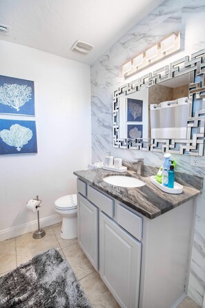 A modern bathroom with a marble sink, a mirror with a geometric frame, and two wall-mounted artwork pieces.