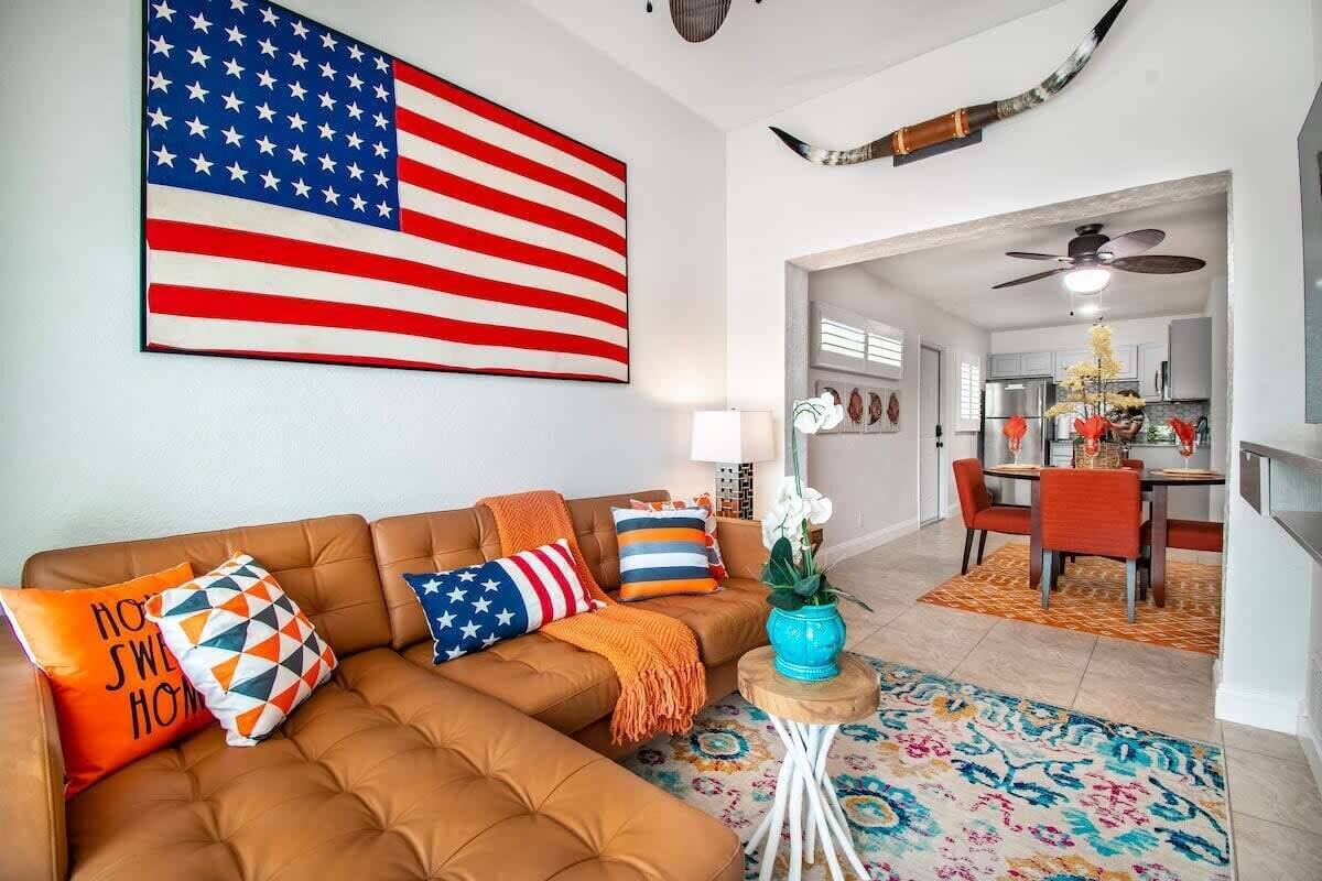 A living room with a brown sectional sofa, a large American flag, and a dining area with orange chairs and a table.