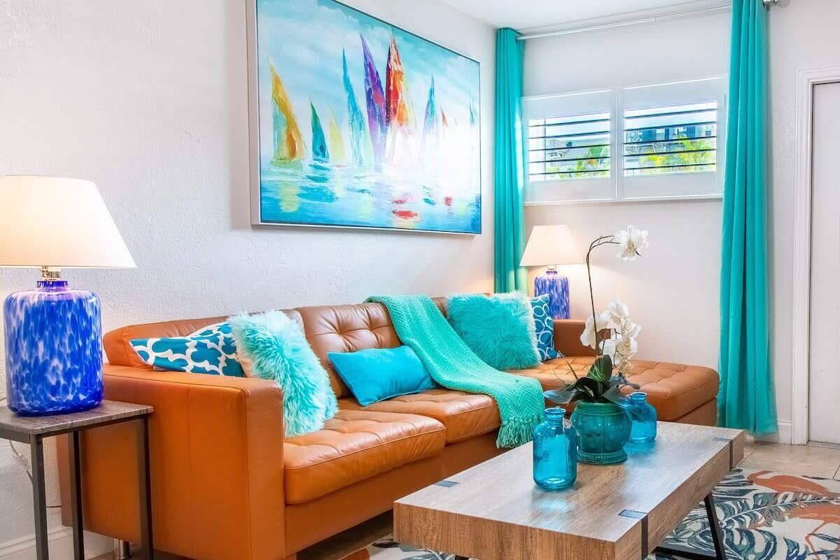 A living room with a brown leather sofa, teal cushions, a painting of sailboats, a window with curtains, and a coffee table with blue vases.