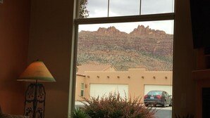 A room with a large window overlooking a mountainous landscape, a table, a lamp, and a couch.