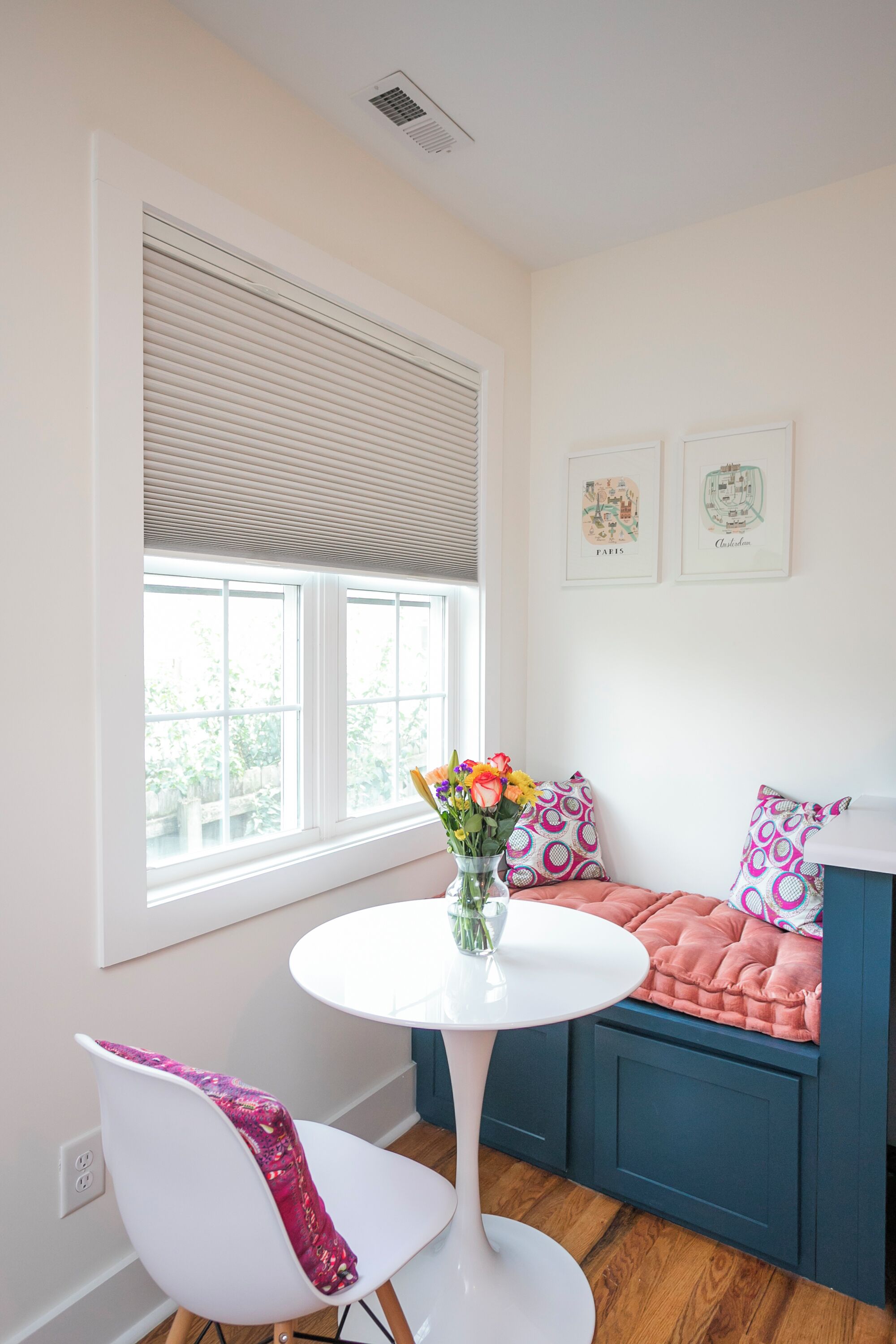 Une pièce avec une table blanche, un canapé bleu, une fenêtre avec des stores et deux tableaux accrochés au mur.
