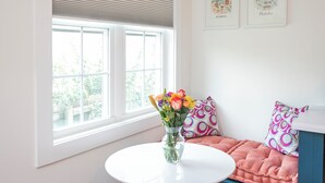 Une pièce avec une table blanche, un canapé bleu, une fenêtre avec des stores et deux tableaux accrochés au mur.