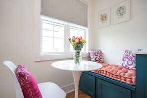 Quarto com uma mesa branca, um banco azul e uma janela com persianas.