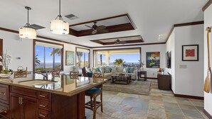 A spacious kitchen with a large island, a dining area with a view, and a living room with a ceiling fan and artwork. - Mauna Kea Waiulaula I201- Stunning Ocean & Golf Views - New Furnishings (Kamuela)