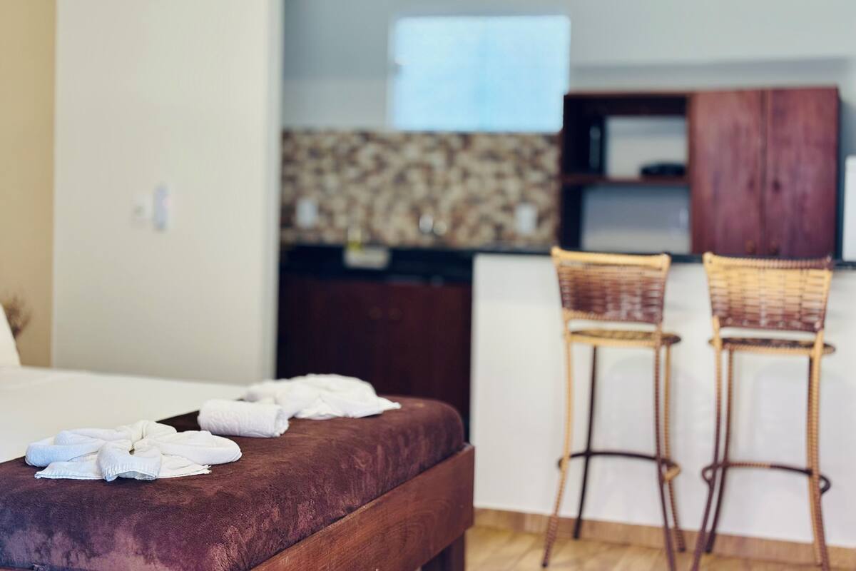 Habitación de hotel con una cama, dos sillas de mimbre, un armario de madera y una kitchenette al fondo.