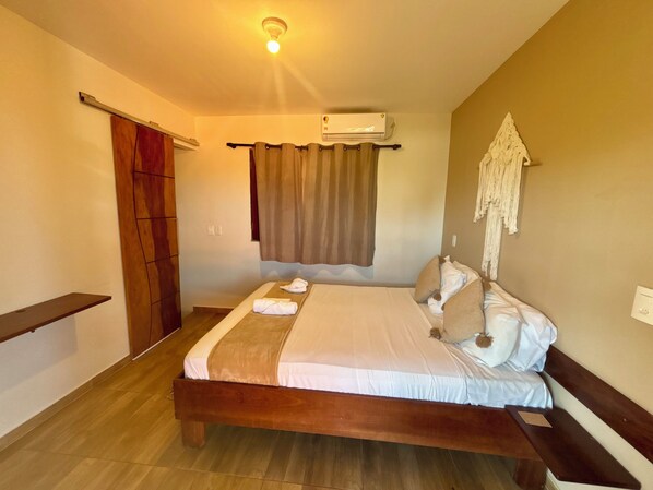 A bedroom with a wooden bed, a ceiling light, an air conditioning unit, a window with curtains, and a wall-mounted macrame piece.