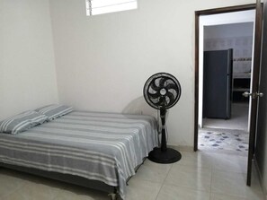 Une petite pièce, peu meublée, avec un lit simple, un ventilateur sur pied et une porte donnant sur une autre pièce.