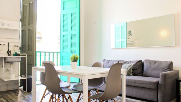 Ein modernes Wohnzimmer mit einem weißen Esstisch, grauen Stühlen und einem grauen Sofa. Ein großes Fenster mit grünen Rollläden, eine Topfpflanze auf dem Tisch und ein Spiegel an der Wand.