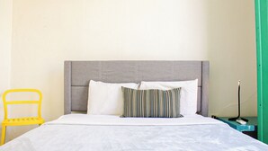 Ein Bett mit grauem Kopfteil, weißen Kissen und einem gestreiften Kissen. Ein gelber Sessel und eine grüne Wand sind ebenfalls zu sehen.