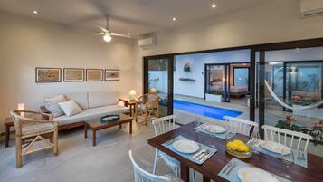 Una sala de estar moderna con un sofá, una mesa de comedor para dos personas y vista a una piscina y zona de estar exterior.